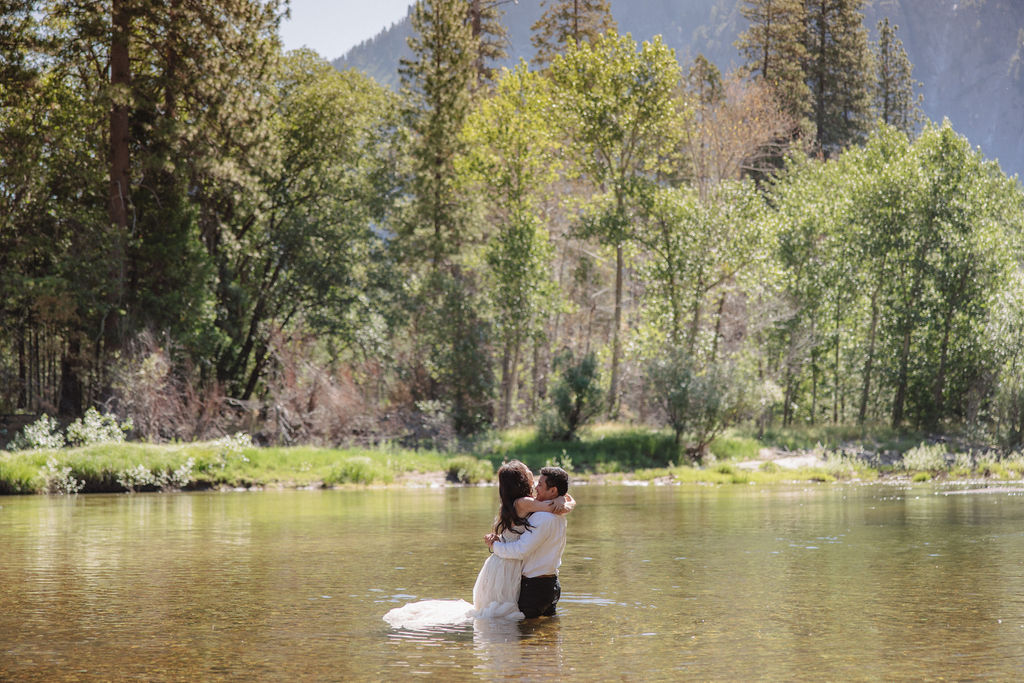 A woman in a white dress and a man in dress clothes wade in a shallow, clear river surrounded by trees and greenery.