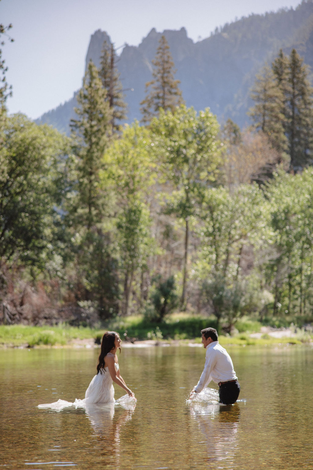 A woman in a white dress and a man in dress clothes wade in a shallow, clear river surrounded by trees and greenery.