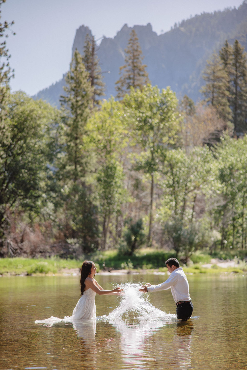 A woman in a white dress and a man in dress clothes wade in a shallow, clear river surrounded by trees and greenery.