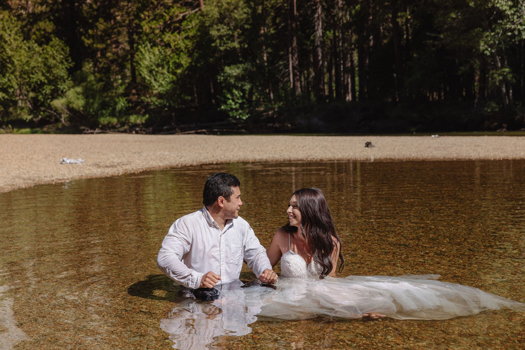 A woman in a white dress and a man in dress clothes wade in a shallow, clear river surrounded by trees and greenery.