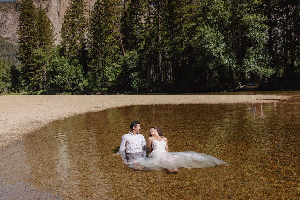 A woman in a white dress and a man in dress clothes wade in a shallow, clear river surrounded by trees and greenery.