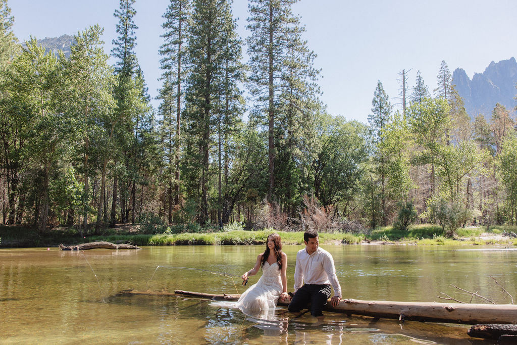 A woman in a white dress and a man in dress clothes wade in a shallow, clear river surrounded by trees and greenery.