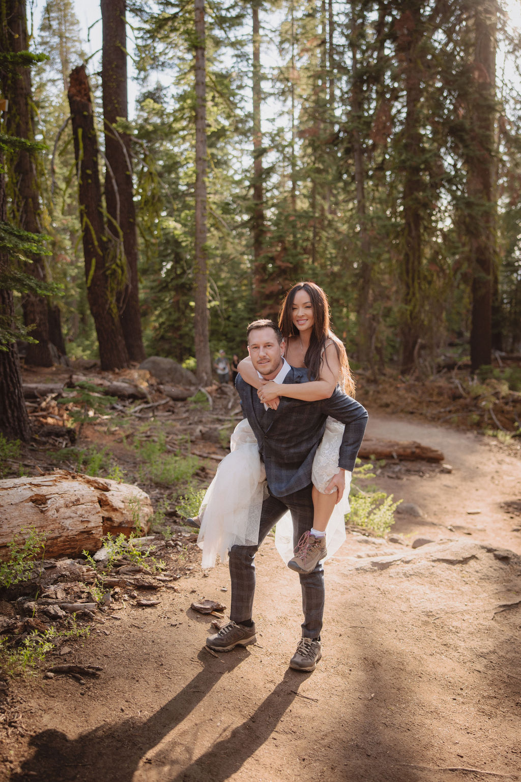 A couple, both wearing backpacks, share a kiss on a forest trail surrounded by trees and foliage for their yosemite elopement