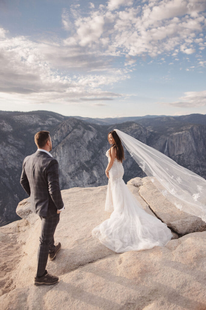 A couple in wedding attire stands on a rocky overlook, facing each other against a backdrop of mountains at sunset.