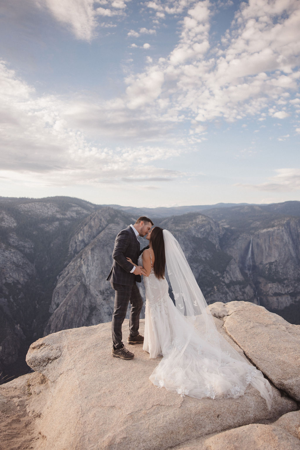 A bride and groom stand together on a cliff edge overlooking a mountainous landscape. The bride is holding the groom's arm and wears a white dress with a long veil, while the groom is in a checked suit for their elopement in yosemite