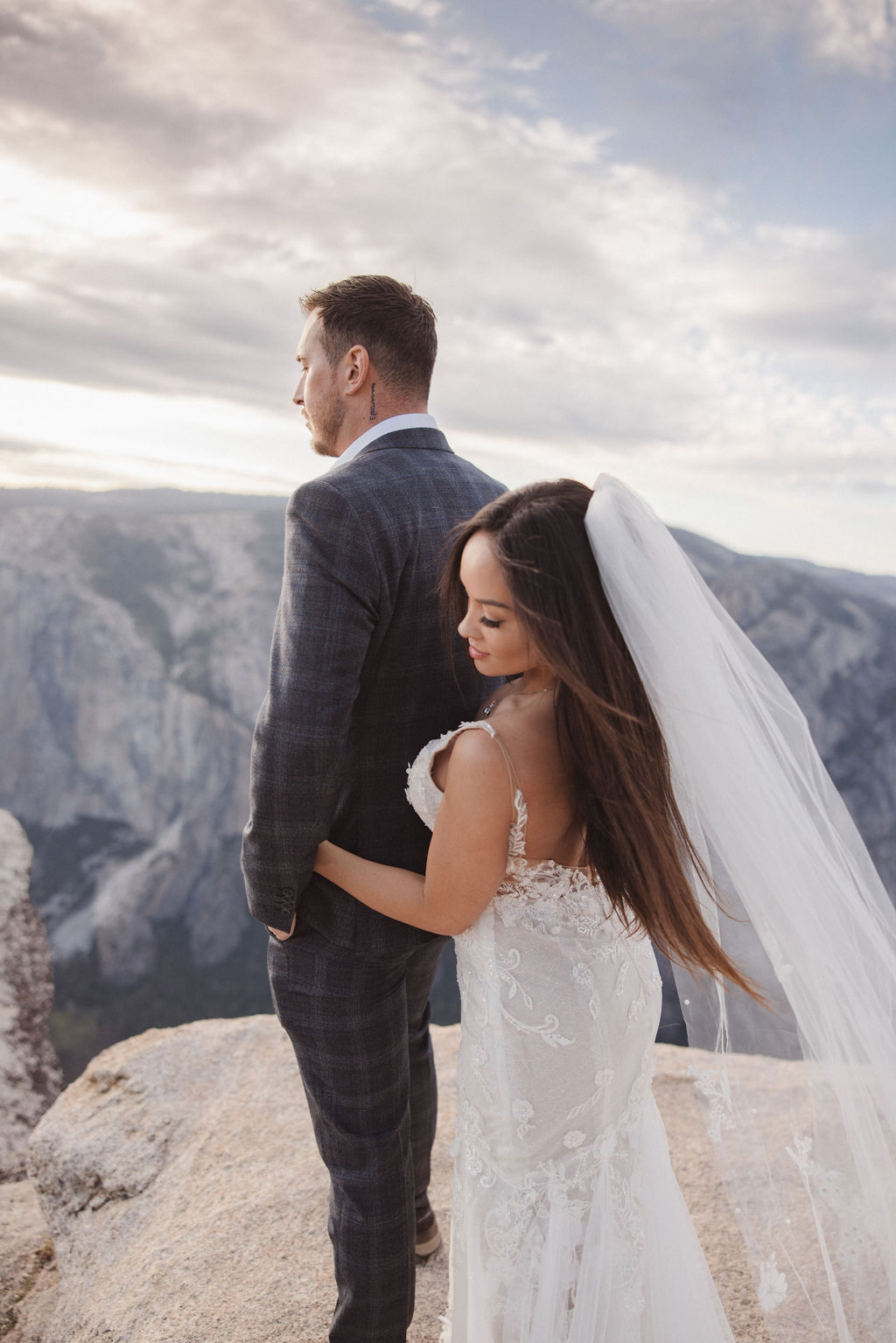 A bride and groom stand together on a cliff edge overlooking a mountainous landscape. The bride is holding the groom's arm and wears a white dress with a long veil, while the groom is in a checked suit for their elopement in yosemite