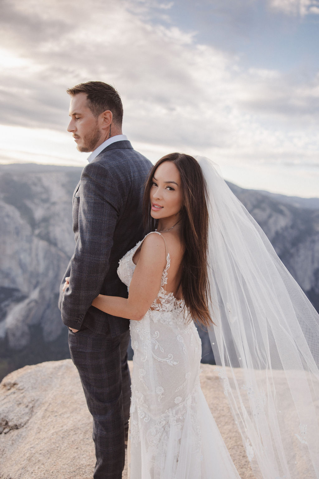 A bride and groom stand together on a cliff edge overlooking a mountainous landscape. The bride is holding the groom's arm and wears a white dress with a long veil, while the groom is in a checked suit for their elopement in yosemite