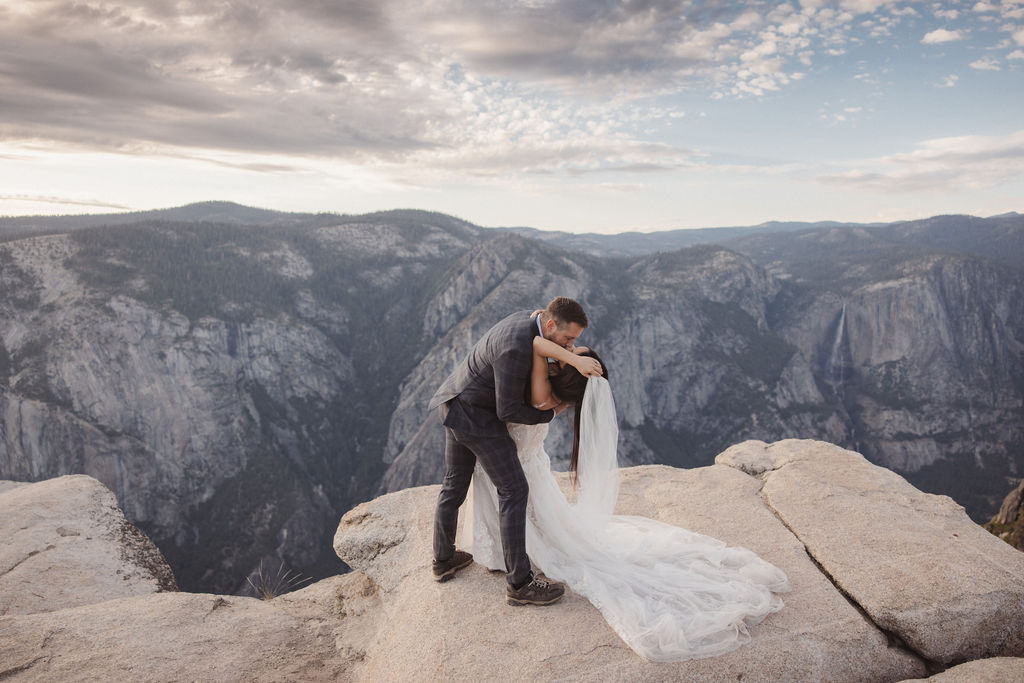 A couple in wedding attire stands on a rocky overlook, facing each other against a backdrop of mountains at sunset.