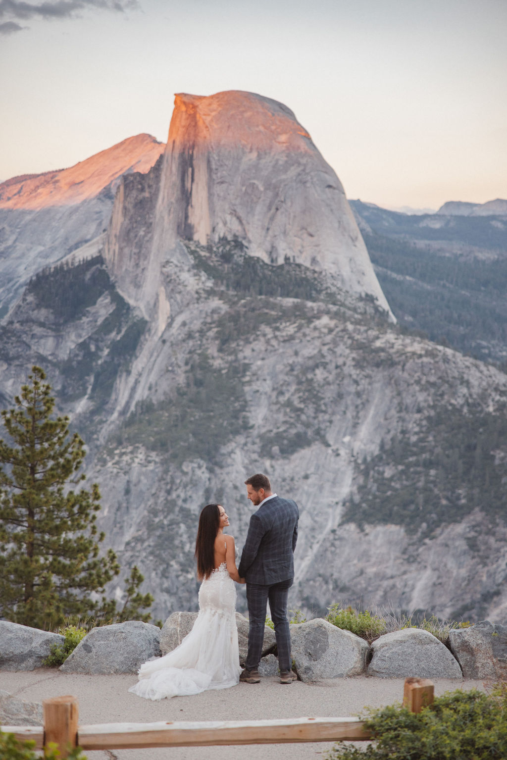 A couple in wedding attire stands on a rocky overlook, facing each other against a backdrop of mountains at sunset.