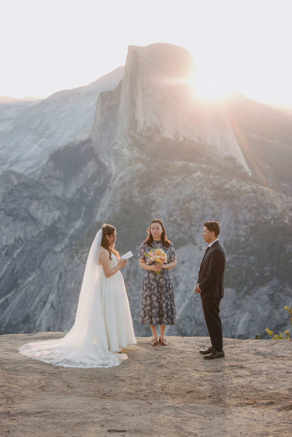 A bride and groom stand facing each other with an officiant between them during an outdoor wedding ceremony with a large mountain in the background in Yosemite