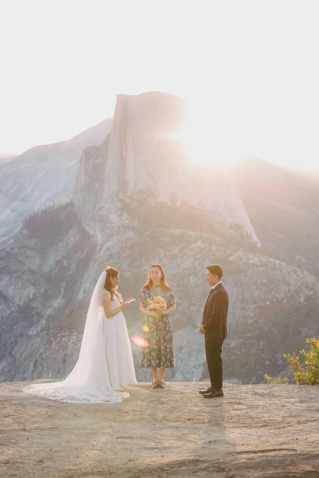 A bride and groom stand facing each other with an officiant between them during an outdoor wedding ceremony with a large mountain in the background in Yosemite 