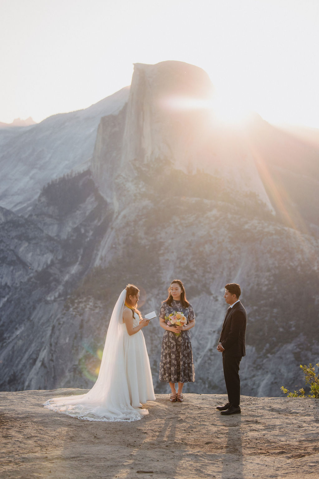 A bride and groom stand facing each other with an officiant between them during an outdoor wedding ceremony with a large mountain in the background in Yosemite 