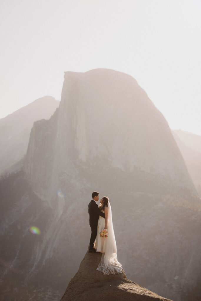 A bride and groom stand together on a rocky cliff with a mountainous landscape in the background, sunlight creating a soft glow for their Yosemite elopement 