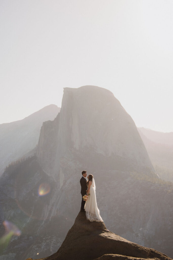 A bride and groom stand together on a rocky cliff with a mountainous landscape in the background, sunlight creating a soft glow for their Yosemite elopement 