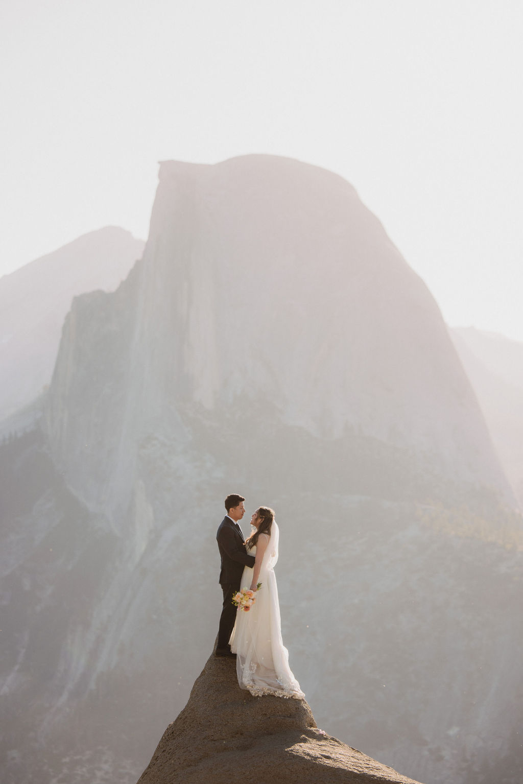 A couple stands on the edge of a cliff with mountainous terrain and a dramatic sky in the background at Glacier point for their elopement at yosemite