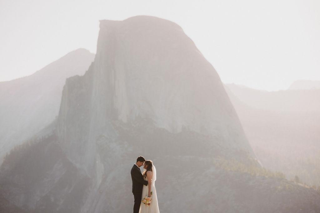 A bride and groom stand together on a rocky cliff with a mountainous landscape in the background, sunlight creating a soft glow for their Yosemite elopement