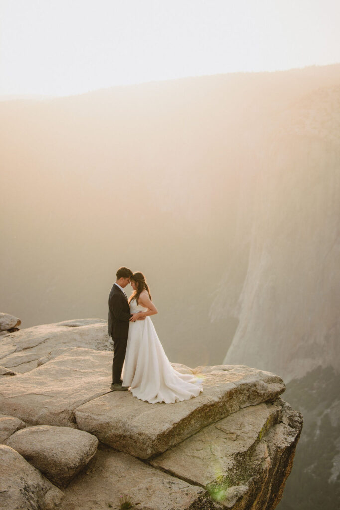 A couple dressed in a wedding gown and suit stand embracing on a rocky cliff edge at sunset for their Yosemite elopement