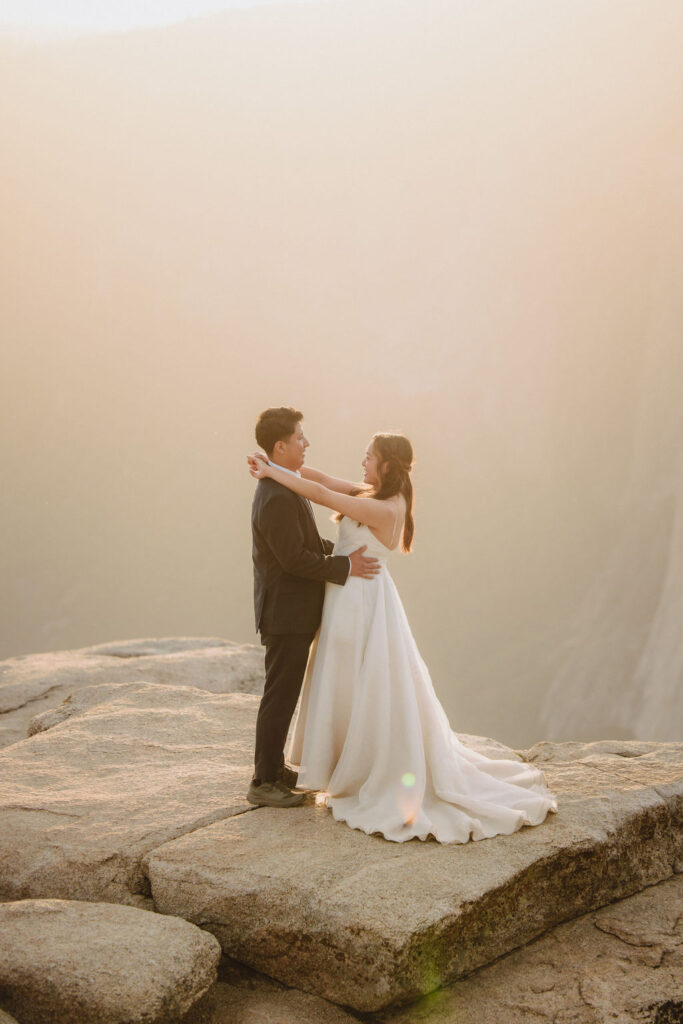 A couple dressed in a wedding gown and suit stand embracing on a rocky cliff edge at sunset for their Yosemite elopement