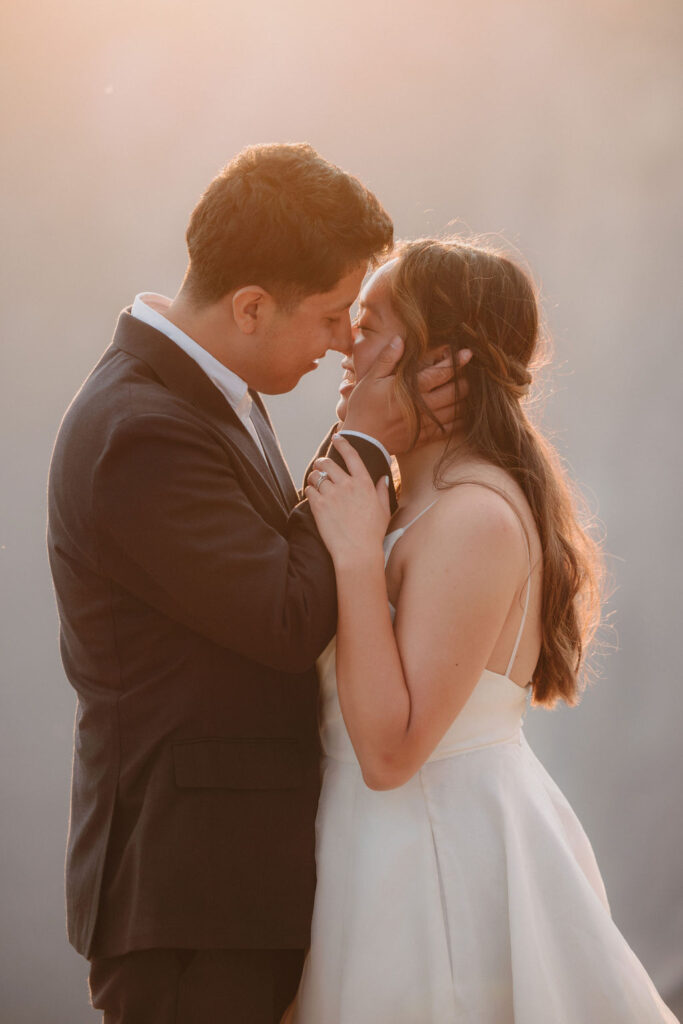 A couple dressed in a wedding gown and suit stand embracing on a rocky cliff edge at sunset for their Yosemite elopement