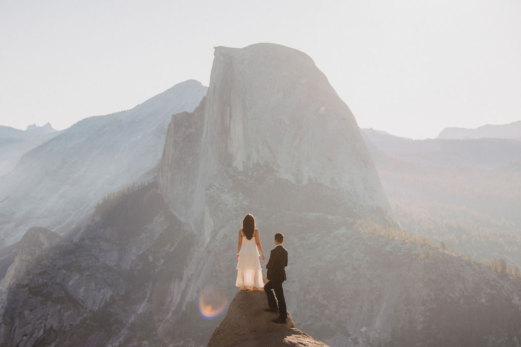 A couple in formal attire stands on a narrow rock ledge with a mountainous backdrop. The man in a suit holds the woman's hand as she wears a long dress, facing each other.