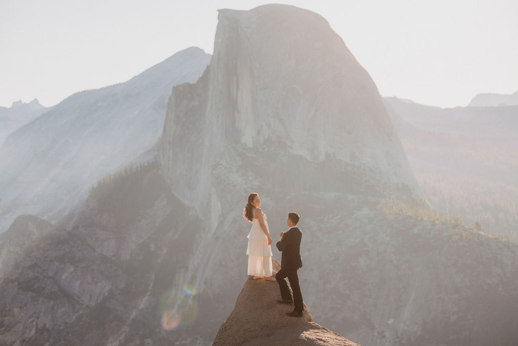 A couple in formal attire stands on a narrow rock ledge with a mountainous backdrop. The man in a suit holds the woman's hand as she wears a long dress, facing each other.