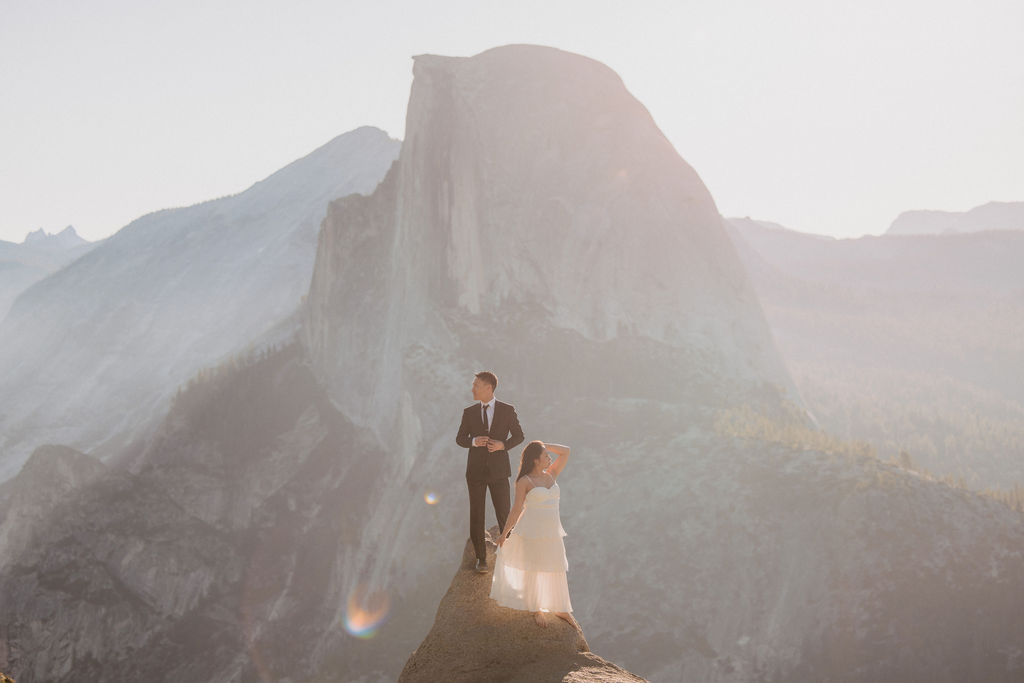 A bride and groom stand on a rocky ledge with a large, misty mountain in the background under a hazy sky for their elopement in Yosemite