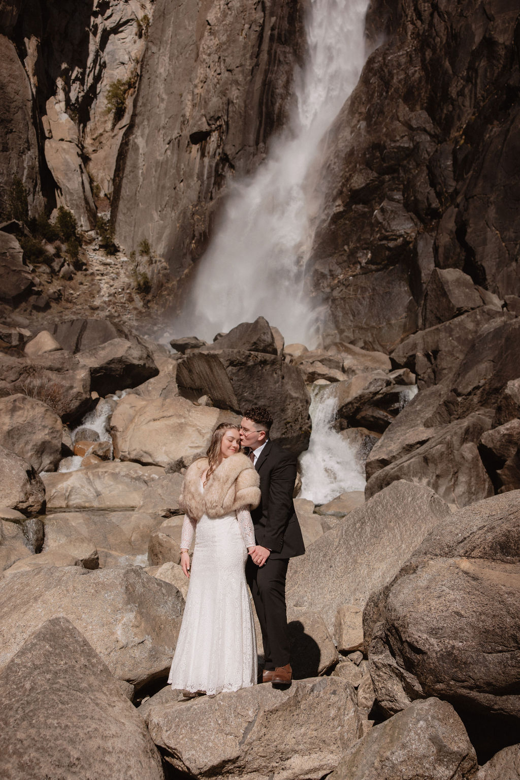 A couple stands on rocks in formal attire with a cascading waterfall behind them at Yosemite falls for their yosemite elopement