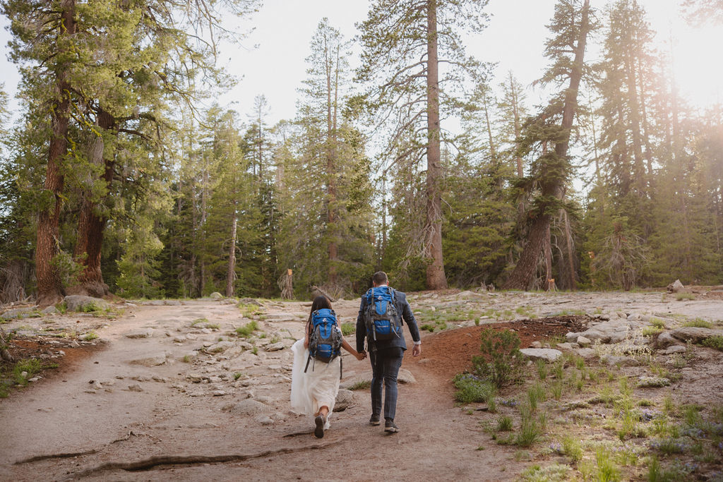 Two people wearing backpacks walk hand-in-hand along a forest trail surrounded by tall pine trees. | Tips for planning a stress free yosemite elopement