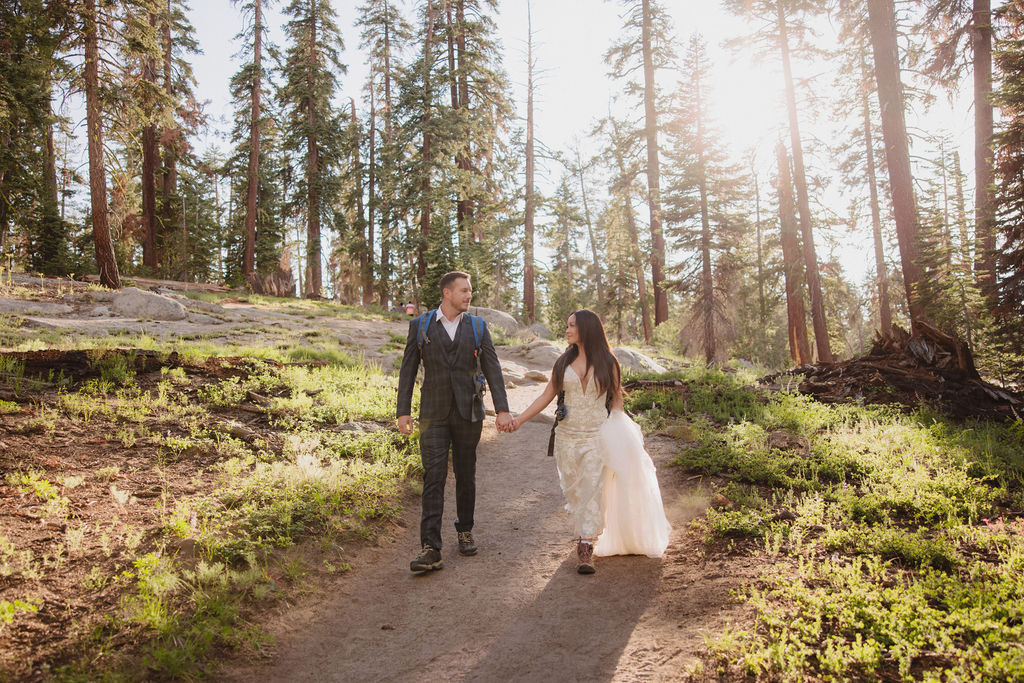 Two people wearing backpacks walk hand-in-hand along a forest trail surrounded by tall pine trees. | Tips for planning a stress free yosemite elopement