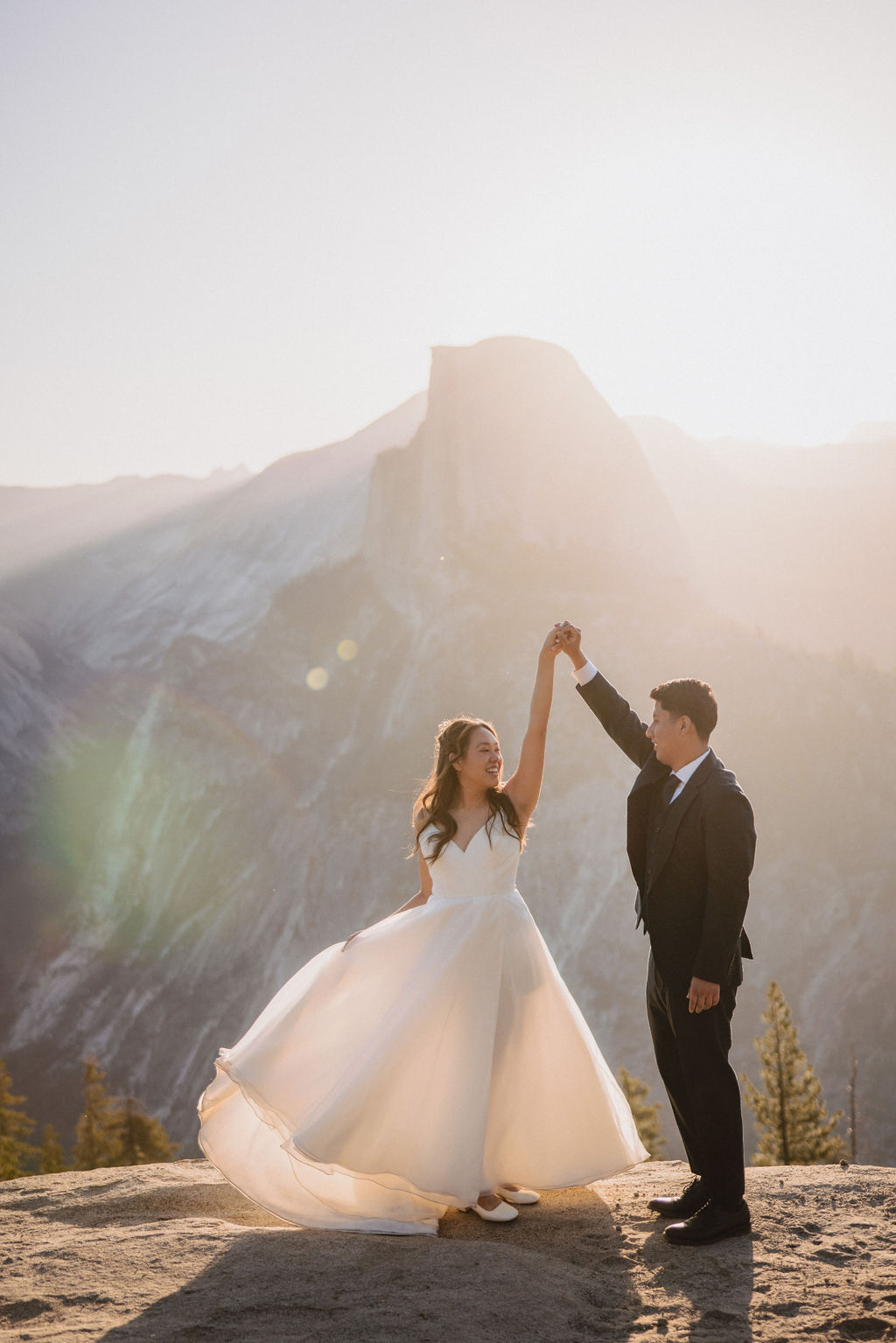 A bride and groom dance on a rock ledge with a mountain backdrop at sunset. The bride is wearing a white dress, and the groom is in a black suit for their Yosemite elopement 