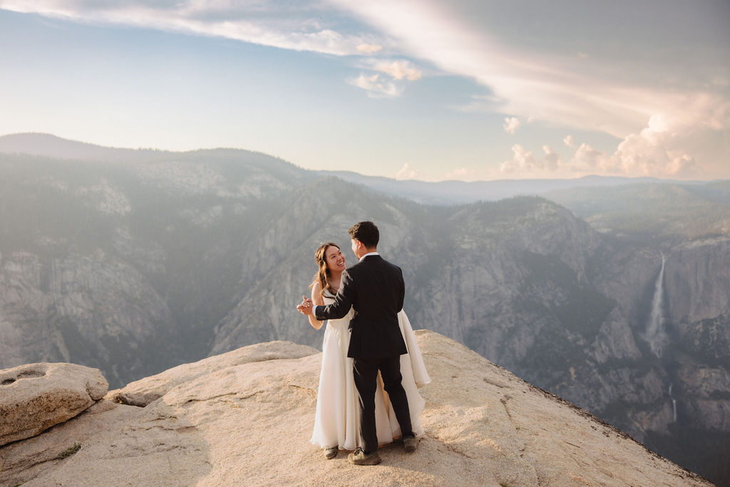 A couple in wedding attire standing on a cliff edge at sunrise or sunset with a mountainous landscape in the background for their yosemite elopement 