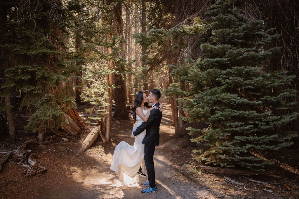 A couple in formal attire, with the woman in a white dress and the man in a suit, pose in an embrace in a forested area with sunlight filtering through the trees. What to pack for a Yosemite elopement
