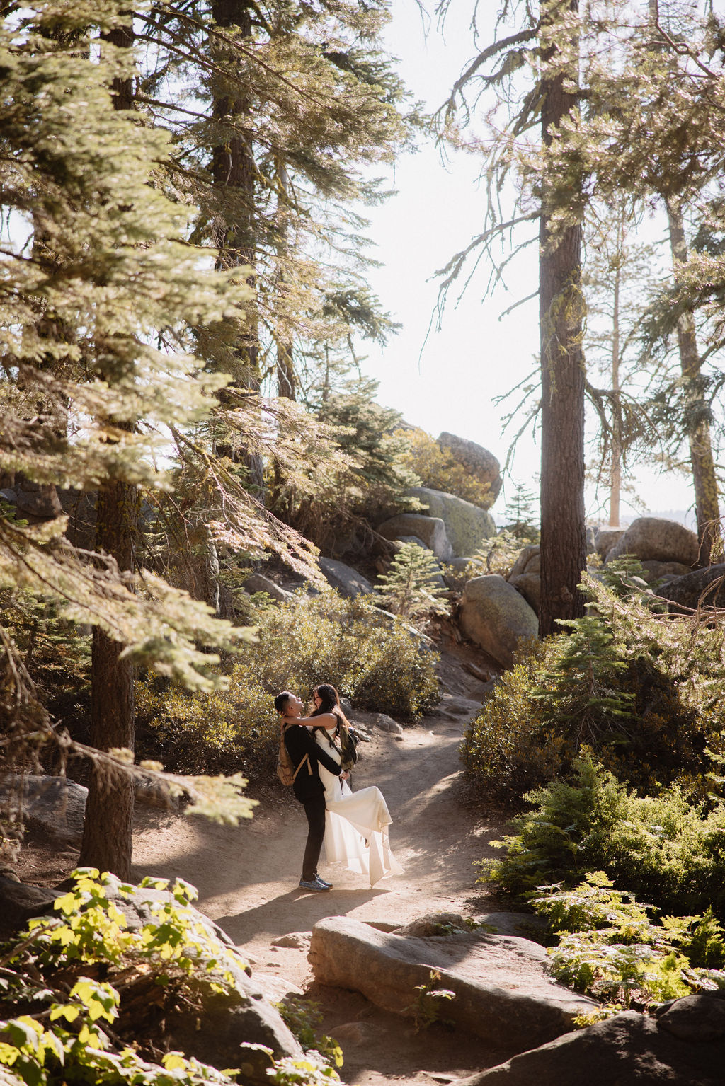 A couple dressed in wedding attire shares an intimate moment in a sunlit forest with tall trees and green foliage. What to pack for a yosemite elopement