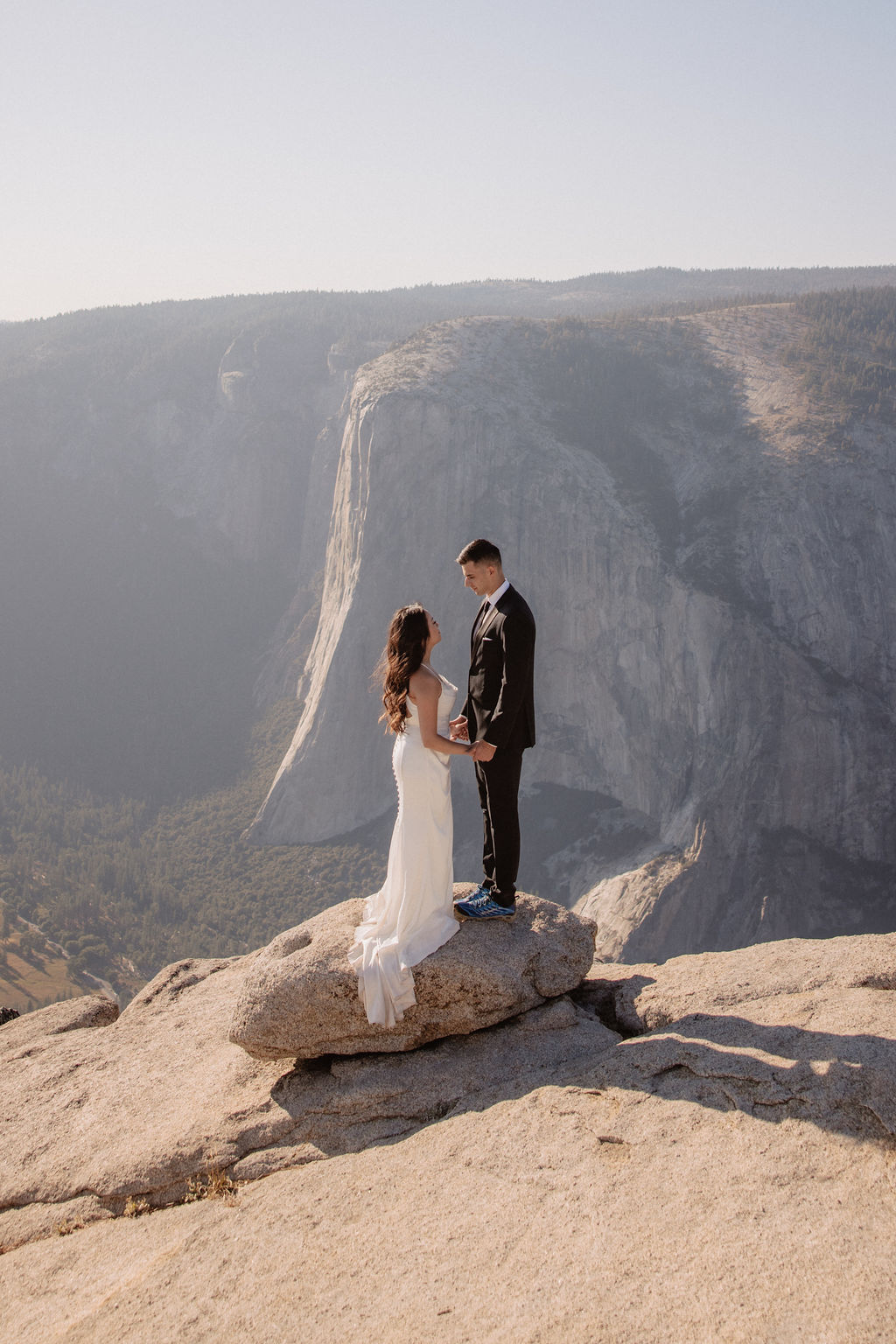 A couple dressed in formal attire stands on a rock at a cliff's edge with a mountainous backdrop. what to pack for a yosemite elopement