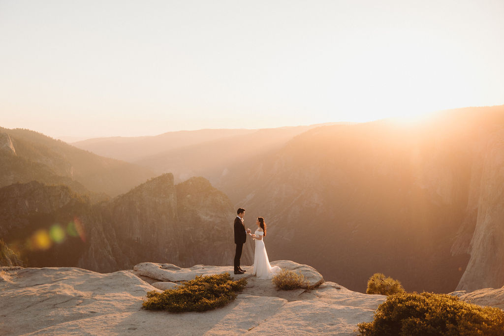 A bride and groom stand holding hands on a rocky cliff at sunset, with hazy mountains and a clear sky in the background.