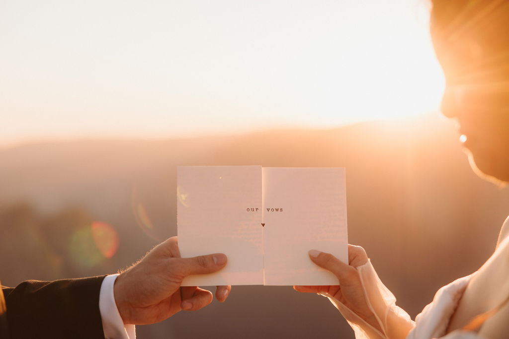 A bride and groom stand holding hands on a rocky cliff at sunset, with hazy mountains and a clear sky in the background.