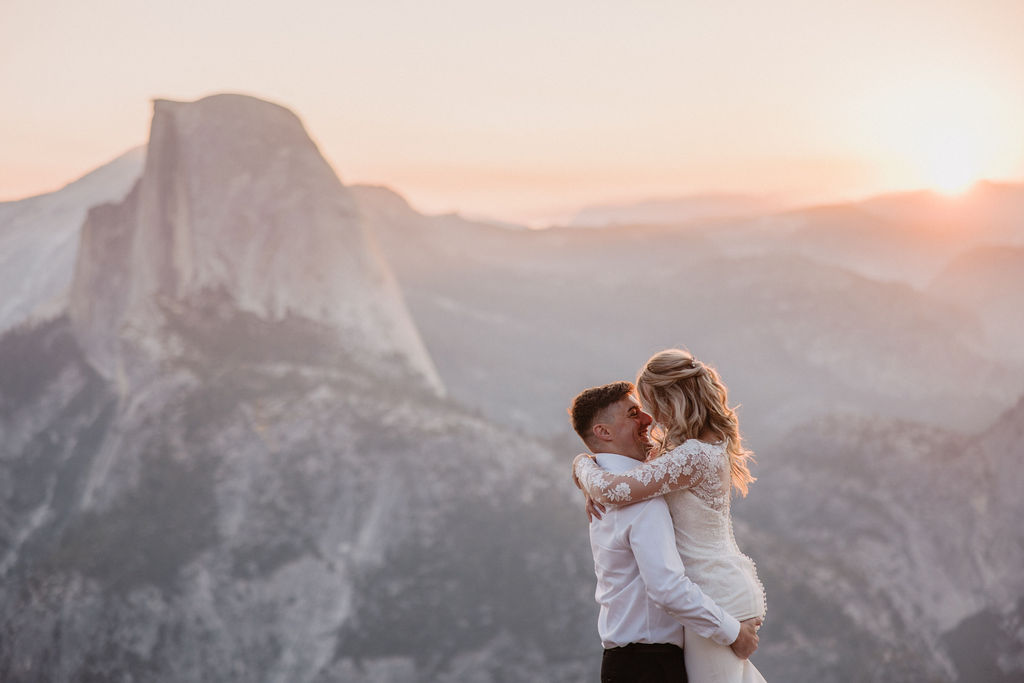 A couple embraces in front of a mountainous landscape at sunset, with a prominent peak in the background. what to pack for a yosemite elopement