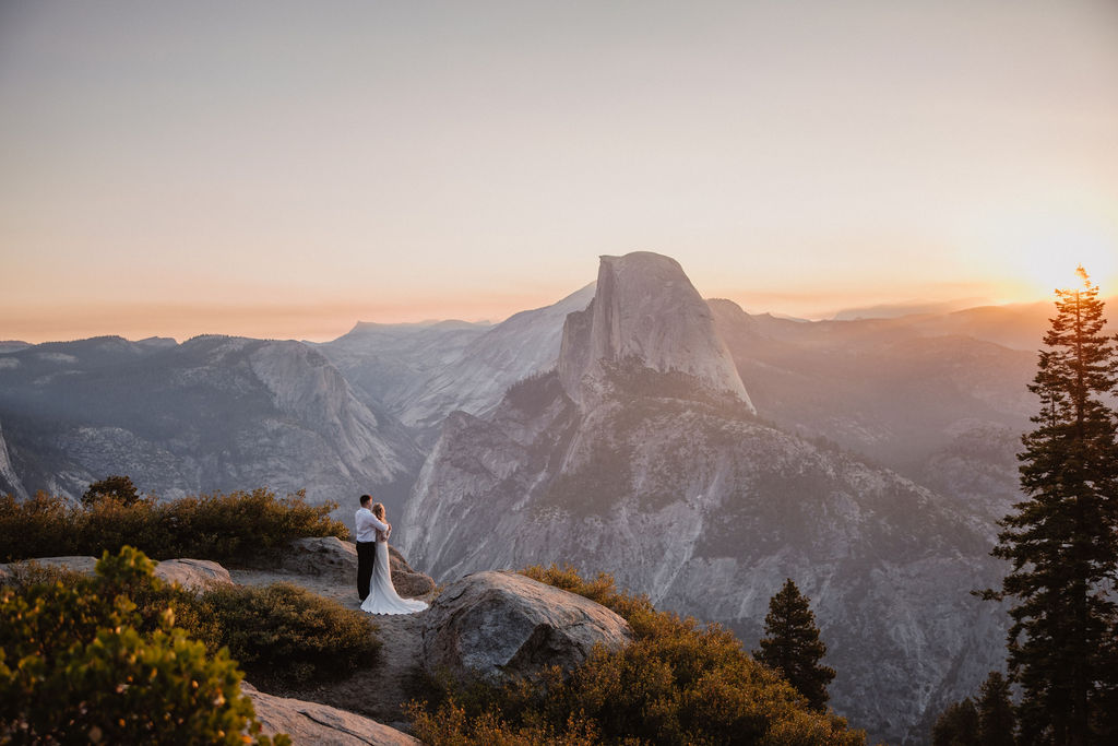 A couple embraces in front of a mountainous landscape at sunset, with a prominent peak in the background. what to pack for a yosemite elopement