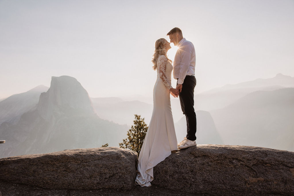 A couple stands hand in hand on a rock, gazing at each other, with a mountainous landscape and a prominent peak in the background A couple stands hand in hand on a rock, gazing at each other, with a mountainous landscape and a prominent peak in the background. What To Pack for a Yosemite Elopement: