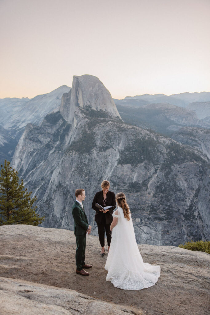 A couple is getting married on a rocky cliff with a scenic mountain landscape in the background; an officiant stands with them. what to pack for a yosemite elopement