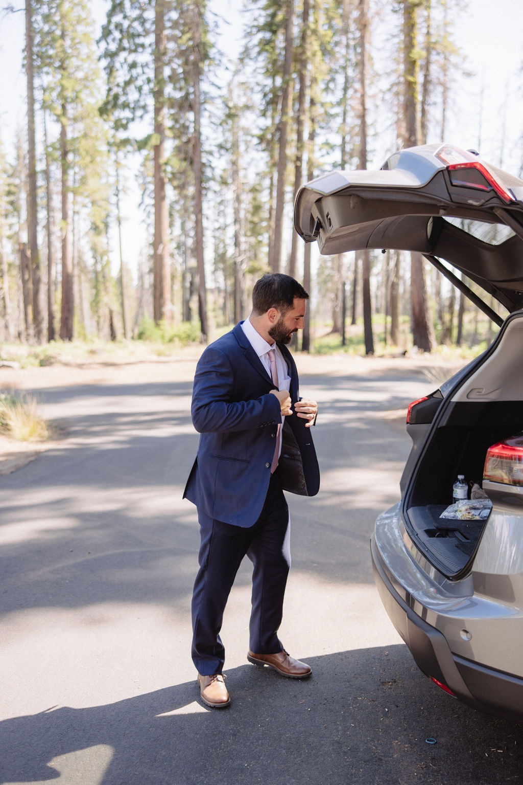 A man in a suit and a woman in casual clothing interact by an open car trunk in a sequoia national park 