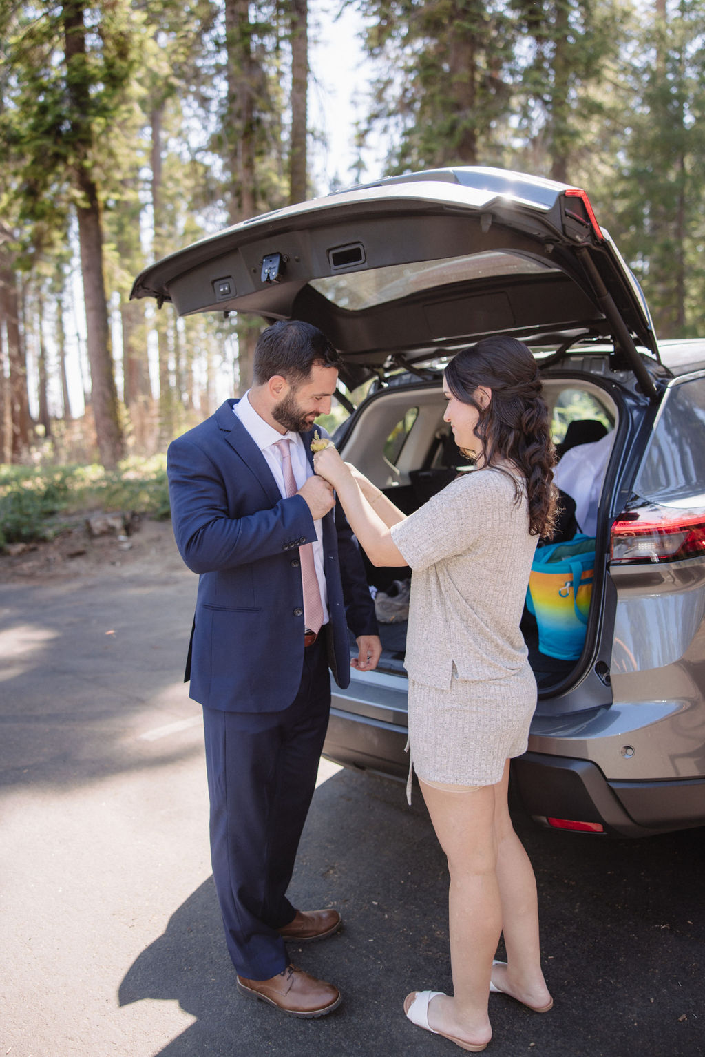 A man in a suit and a woman in casual clothing interact by an open car trunk in a sequoia national park 