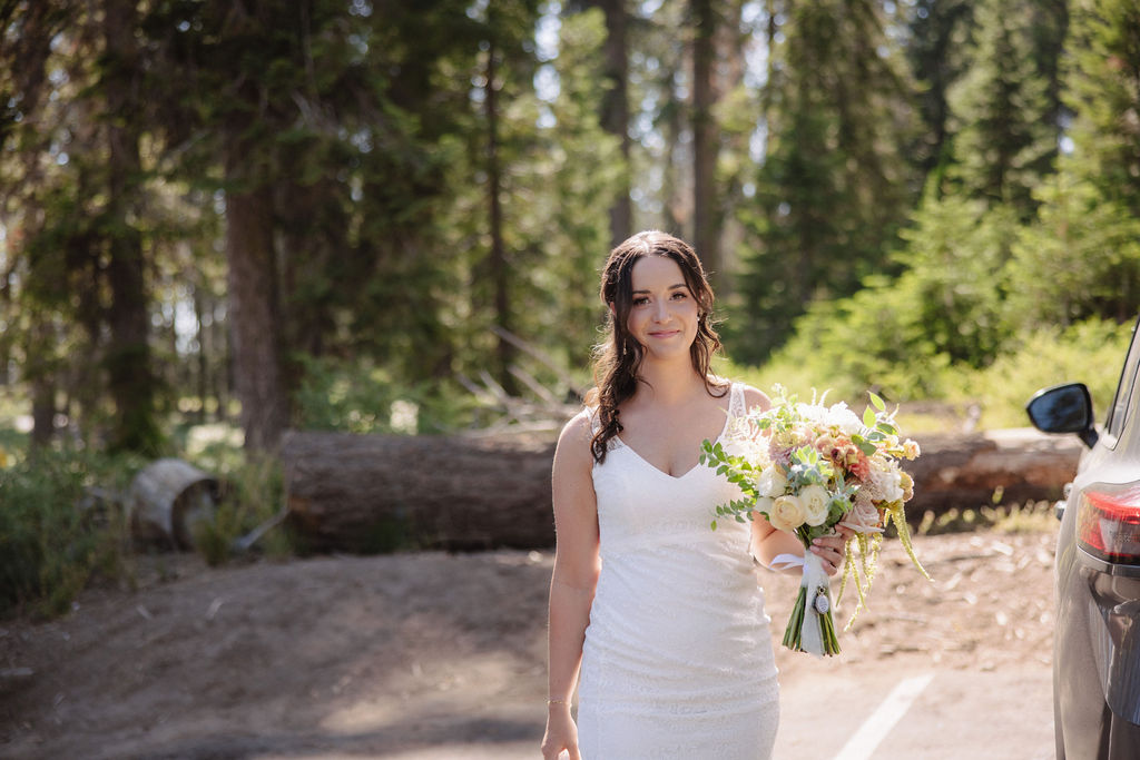 A woman in a white dress holds a bouquet of flowers and stands in a wooded area next to a parked car.