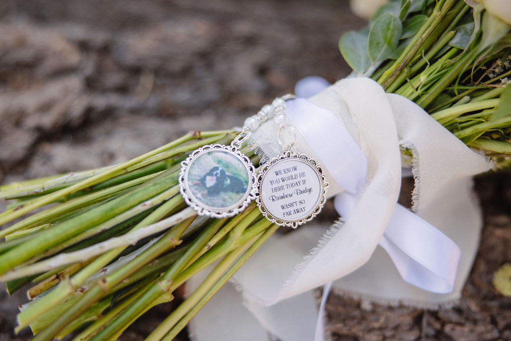 Close-up of two silver lockets attached to a bouquet of flowers, one with a blue gemstone and the other with an engraved message on a white ribbon. The background is a rough, textured surface.