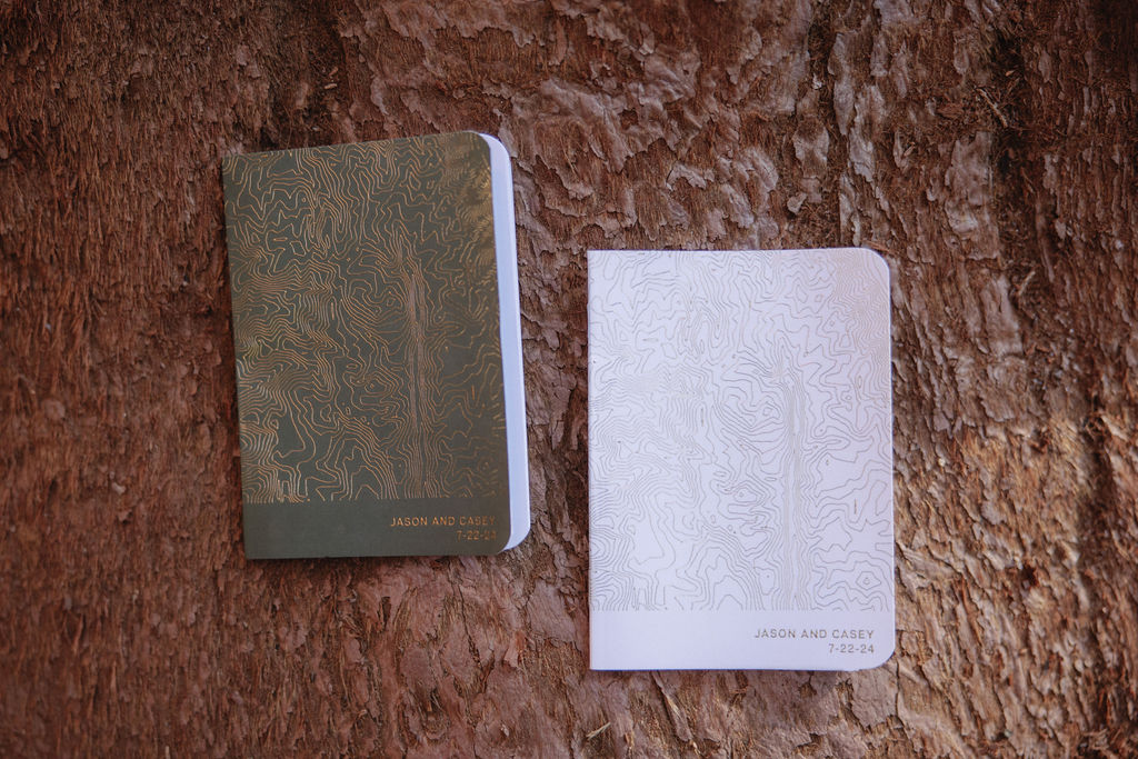 Two notebooks placed on a textured brown surface. The left notebook has a green cover with a golden design, and the right notebook has a white cover with a subtle design.