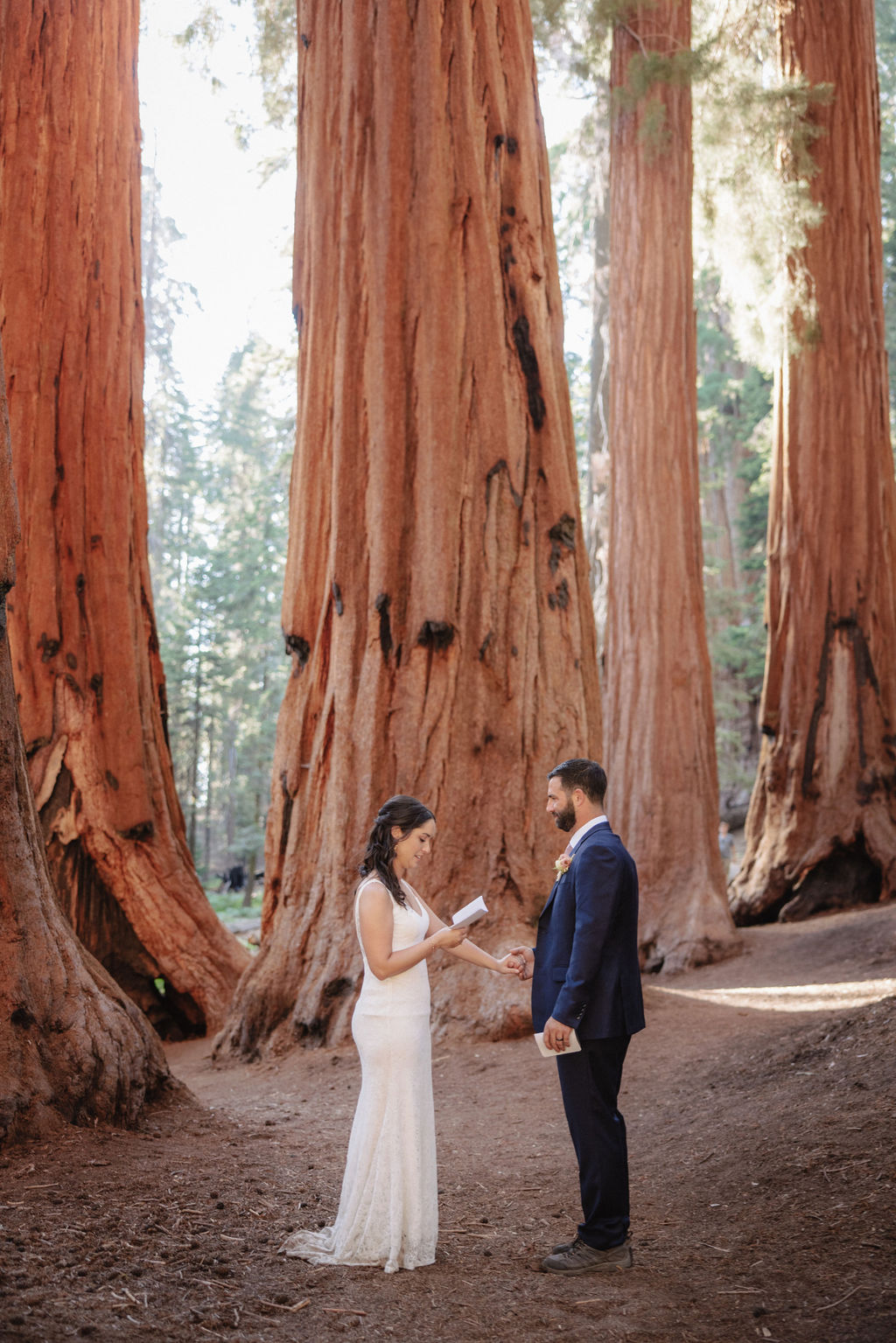 A bride and groom exchange vows in a forest surrounded by giant redwood trees. The bride holds a vow book in her hands, and the groom is looking at her attentively.