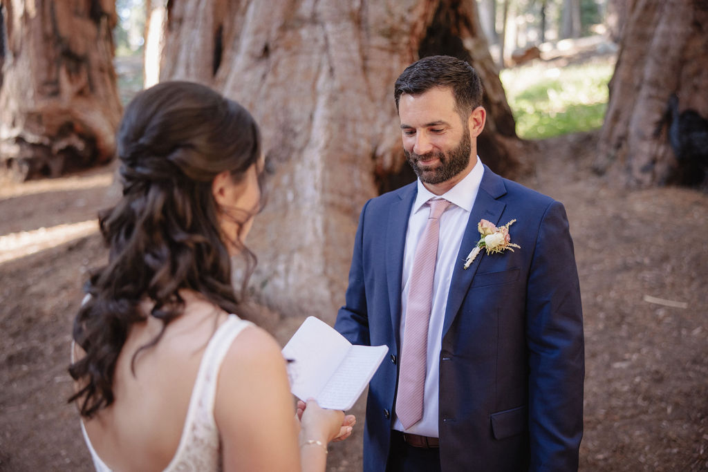 A bride and groom exchange vows in a forest surrounded by giant redwood trees. The bride holds a vow book in her hands, and the groom is looking at her attentively.