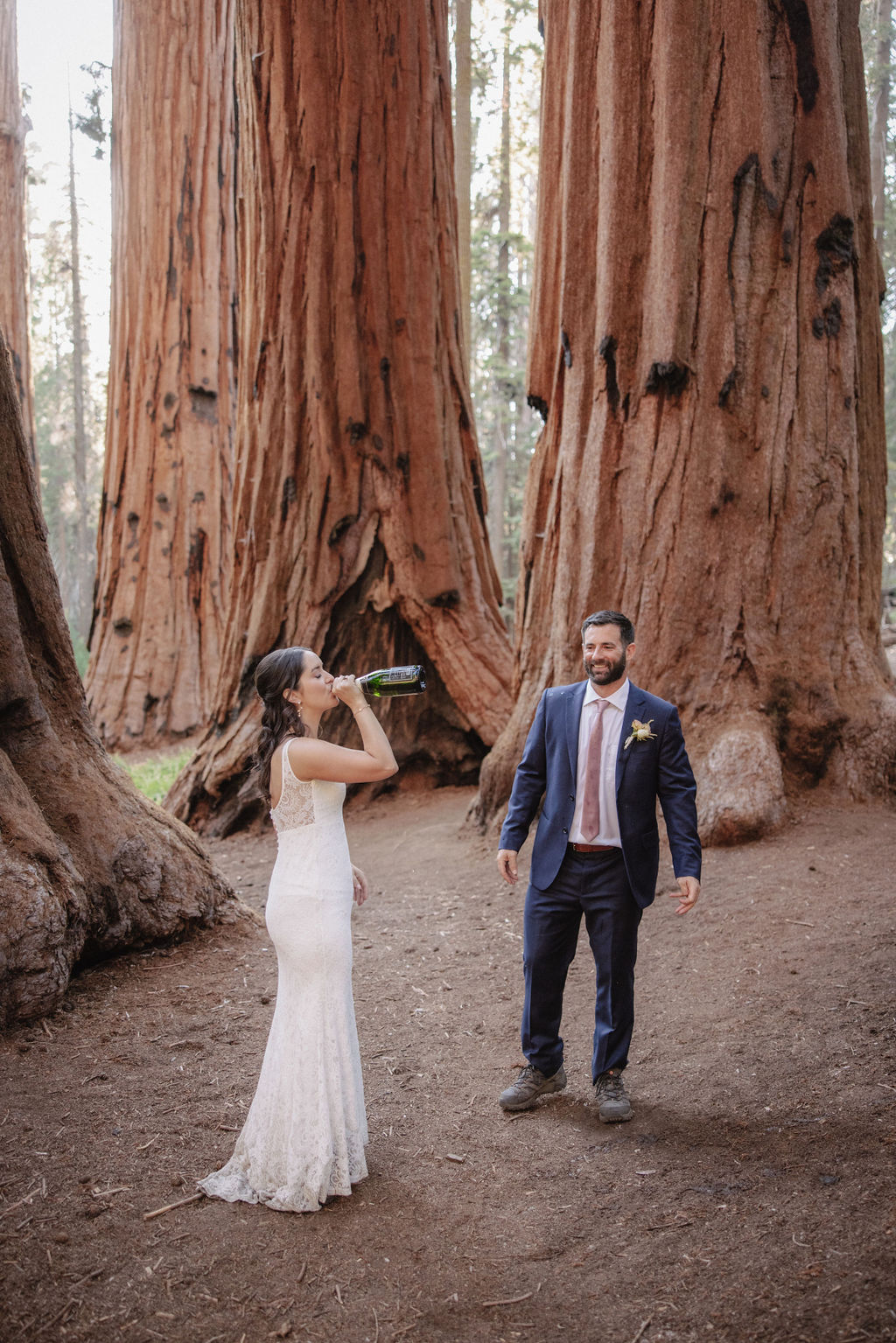 A couple in formal attire stands in a forest of large trees while the man pops a bottle of champagne, causing a spray of bubbles