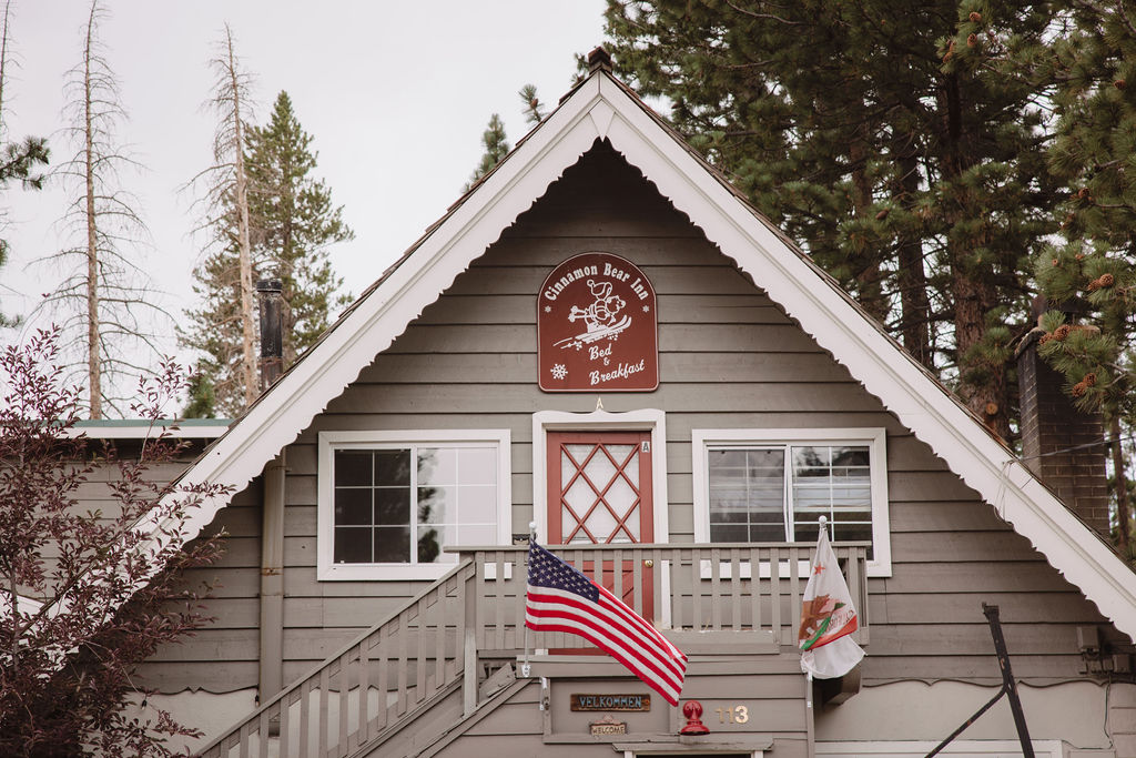 A two-story wooden building with a triangular roof and brown exterior. An American flag and a California state flag hang beside the entrance, with a small sign near the peak of the roof.