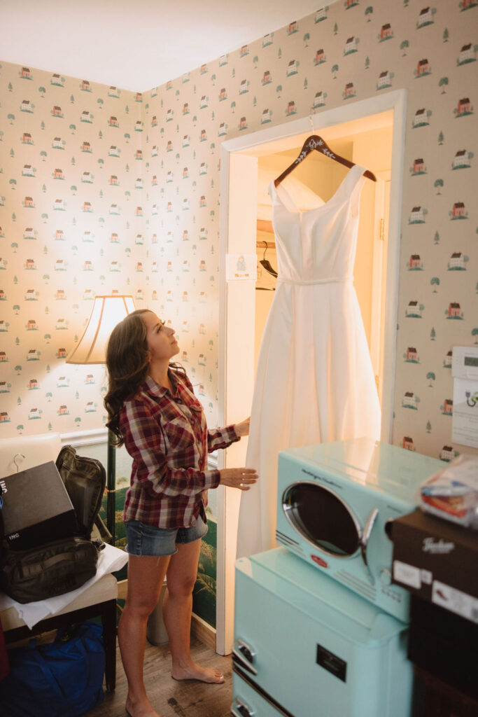 A woman in a plaid shirt and shorts looks up at a white dress hanging in a well-lit room with vintage wallpaper and a turquoise dresser.